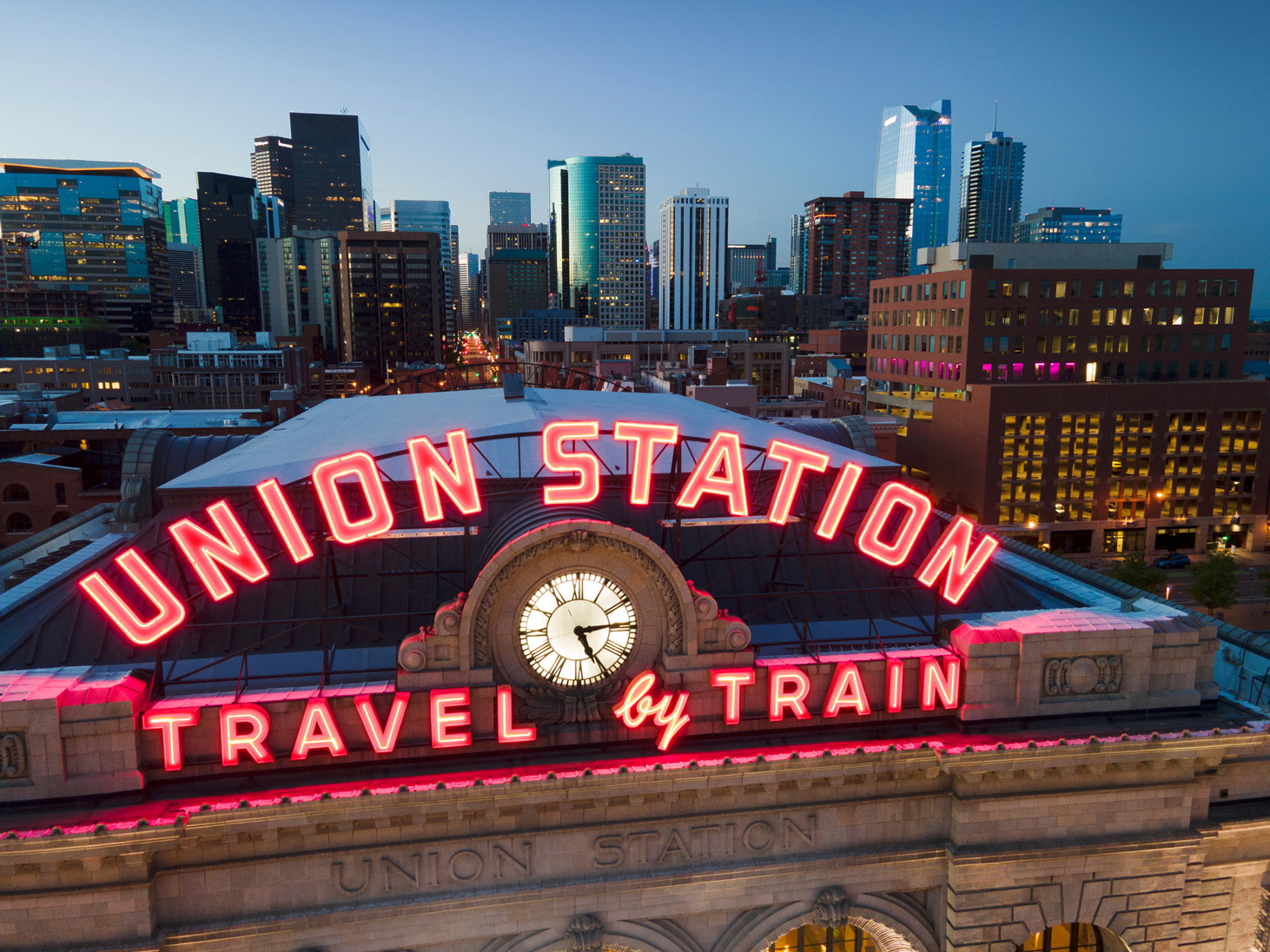 Union Station in evening with neon sign that says travel by train, Denver city skyline behind it